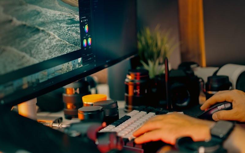 Hands at a workstation editing an image, with cameras and lenses nearby